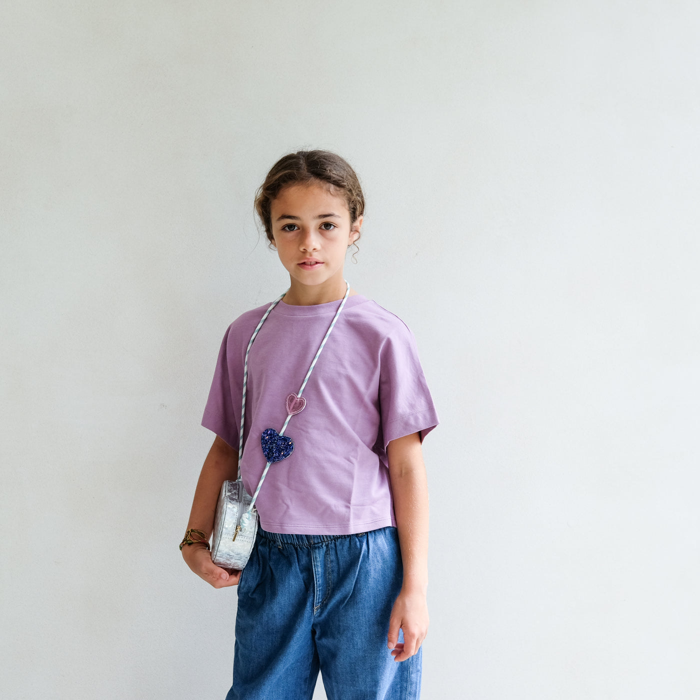 Young girl wearing a purple shirt and blue jeans wearing a silver heart cross body bag against a plain background