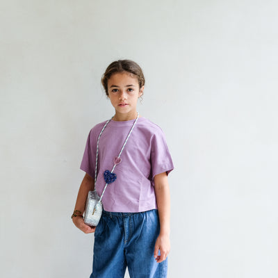 Young girl wearing a purple shirt and blue jeans wearing a silver heart cross body bag against a plain background