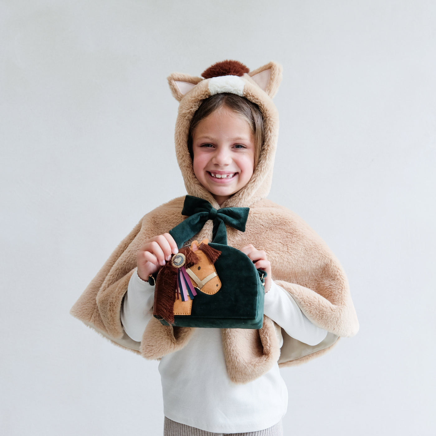 Child wearing a faux fur horse hooded dressing up cape holding a novelty horse bag against a plain background
