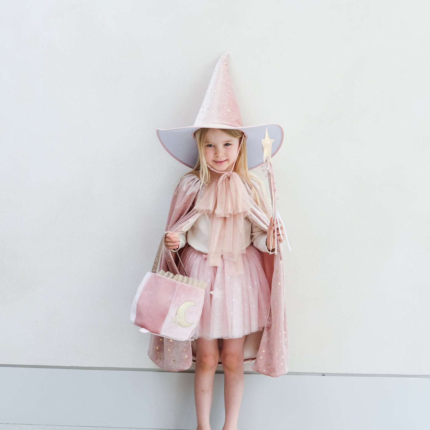 Young girl in a pink outfit with a wide-brimmed pink velvet hat and cape and matching wand, holding a pretty pink cauldron shape trick or treat bag