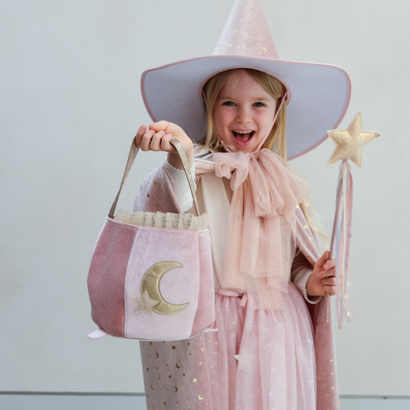 Child in a pink witch costume with a pink hat, bag, and wand against a light background