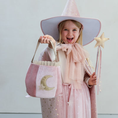 Child in a pink witch costume with a pink hat, bag, and wand against a light background