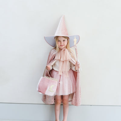 Young girl in a pink outfit with a wide-brimmed pink velvet hat and cape and matching wand, holding a pretty pink cauldron shape trick or treat bag