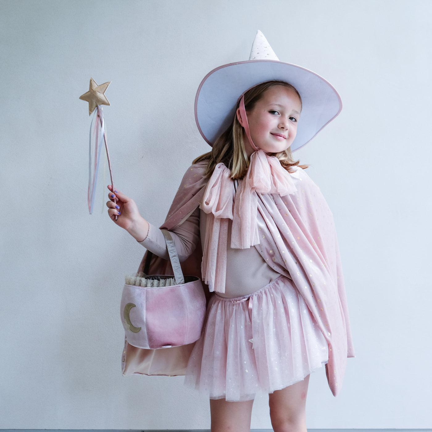 Young girl in a pink outfit with a wide-brimmed pink velvet hat and cape and matching wand, holding a pretty pink cauldron shape trick or treat bag