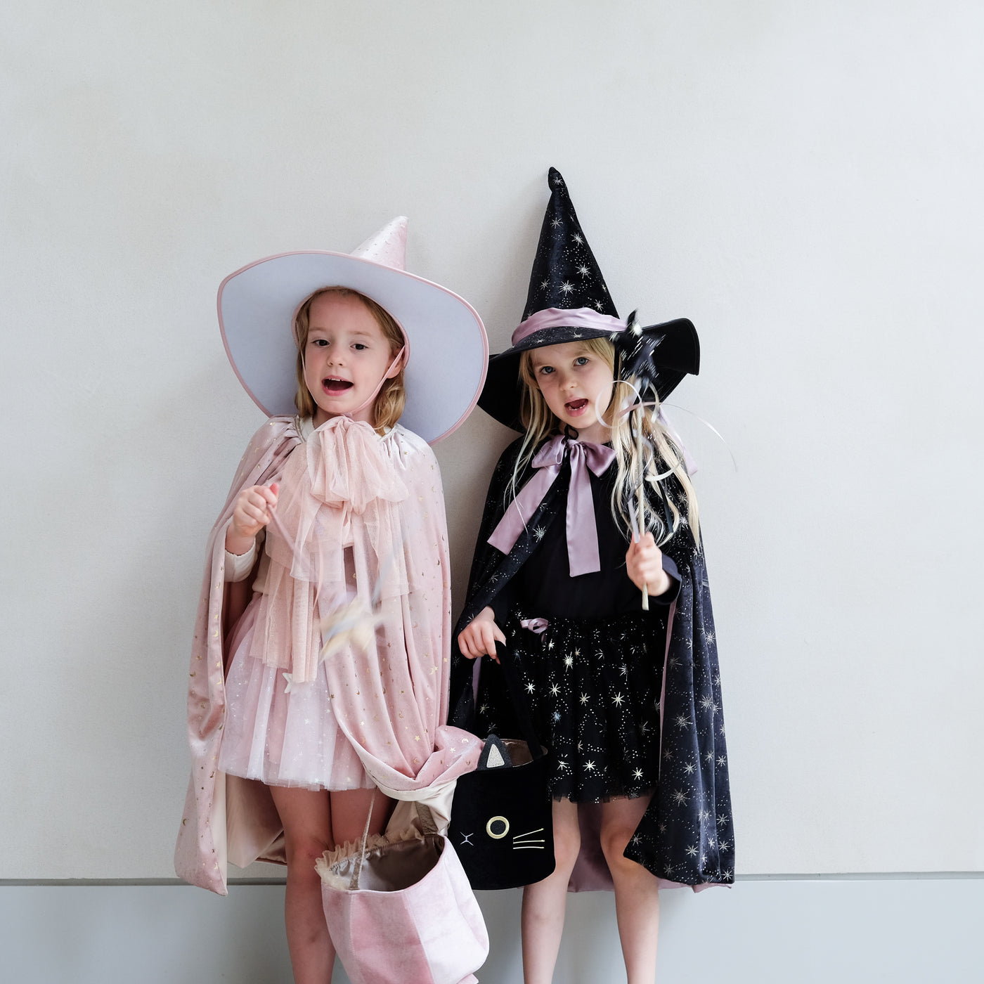 Two children in velvet Halloween costumes, one in a pink witch costume and the other in a black witch costume complete with hats and trick or treat bags