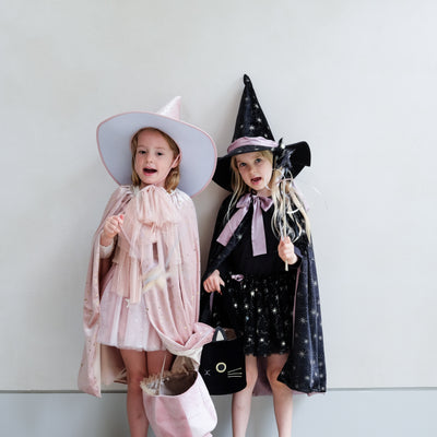 Two children in velvet Halloween costumes, one in a pink witch costume and the other in a black witch costume complete with hats and trick or treat bags