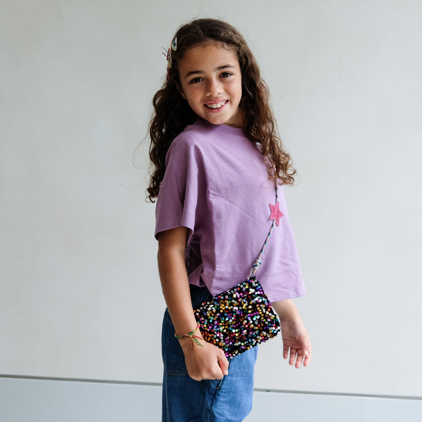 Young girl wearing a purple shirt and blue jeans, holding a colourful sequin bag against a plain background