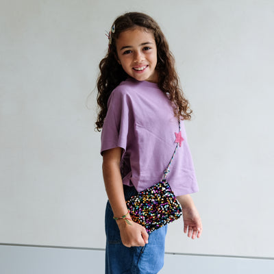 Young girl wearing a purple shirt and blue jeans, holding a colourful sequin bag against a plain background