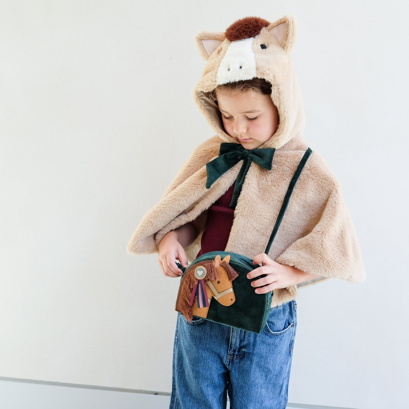 Child wearing a horse-themed cape with a hood and holding a matching bag with horse design against a white background
