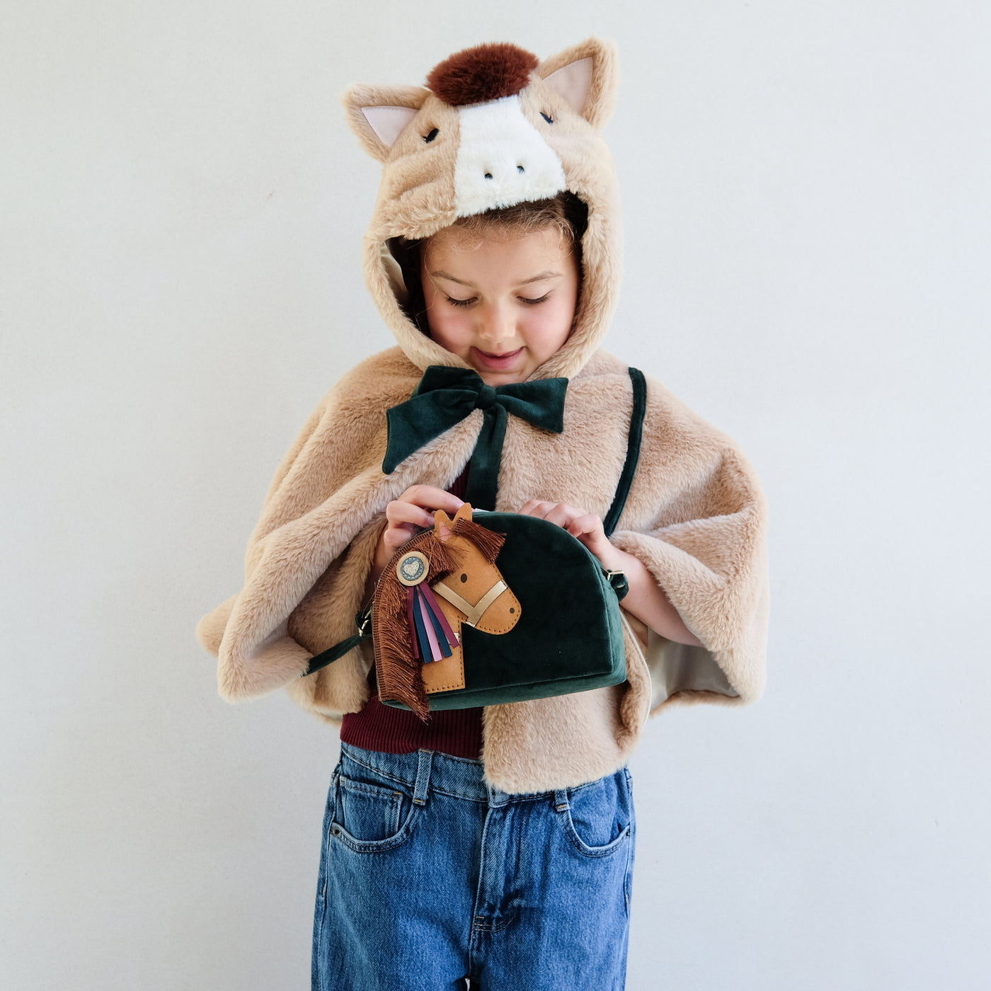 Child wearing a faux fur horse hooded dressing up cape holding a horse novelty cross body bag against a plain background