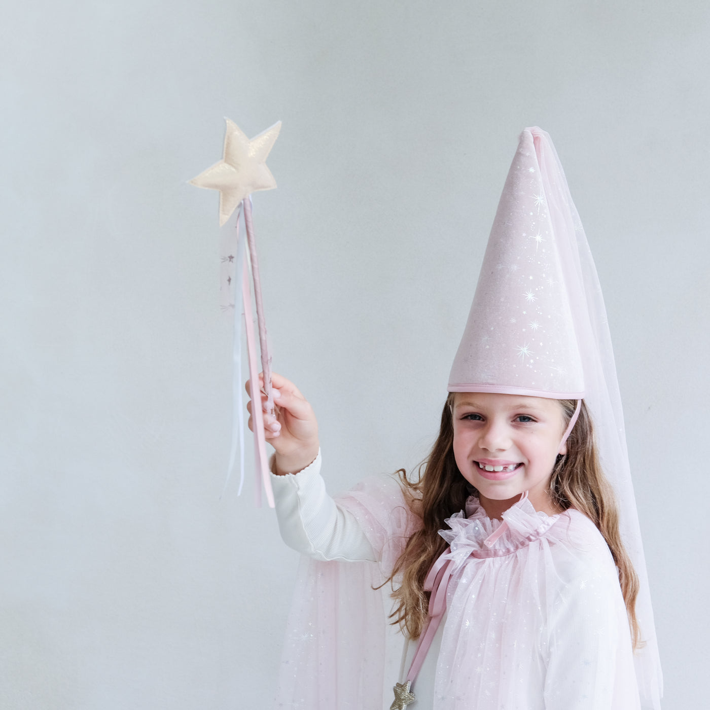 Girl waving a pink star wand wearing a pink princess hat, and pink shimmer cape against a white background