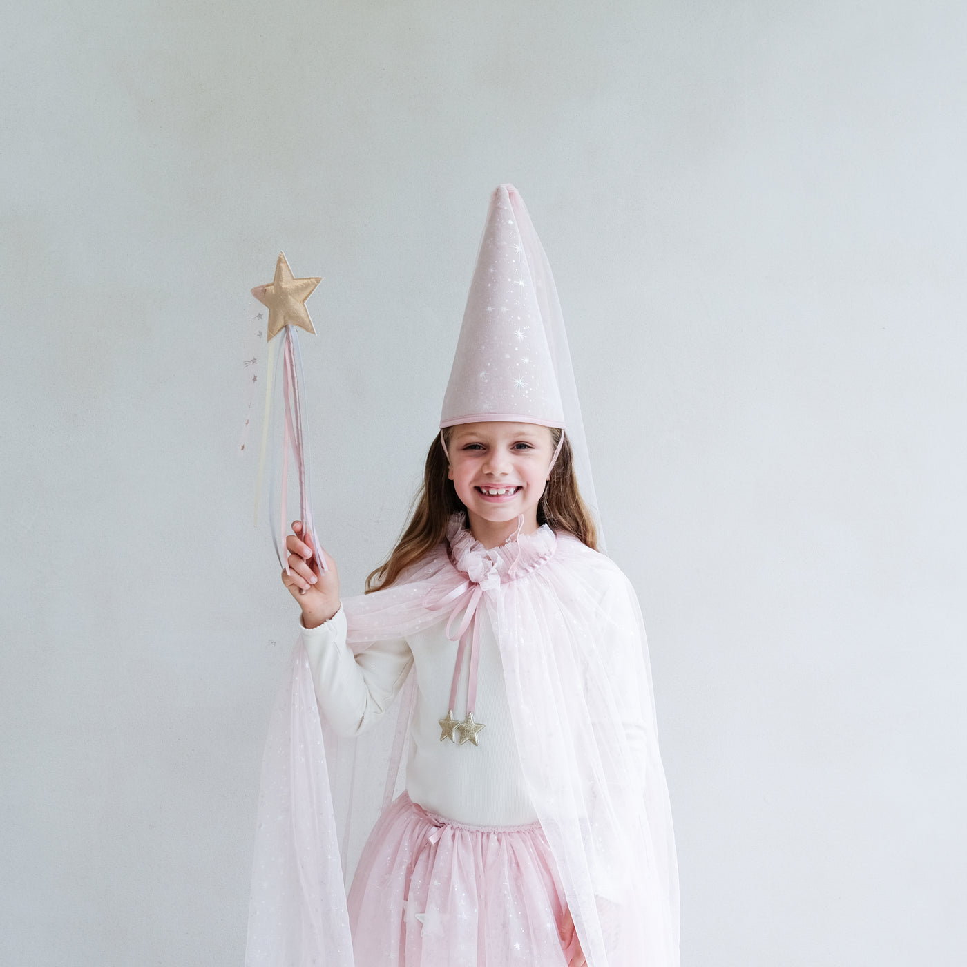 Young girl wearing a full pink shimmer dressing up costume in pink with wand, princess hat, and matching tutu and cape against a white background