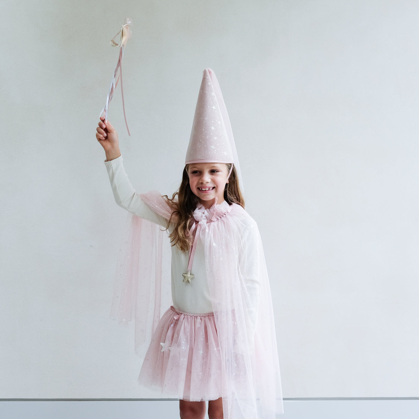 Young girl wearing a full pink shimmer dressing up costume in pink with wand, princess hat, and matching tutu and cape against a white background