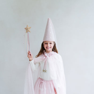 Young girl wearing a full pink shimmer dressing up costume in pink with wand, princess hat, and matching tutu and cape against a white background