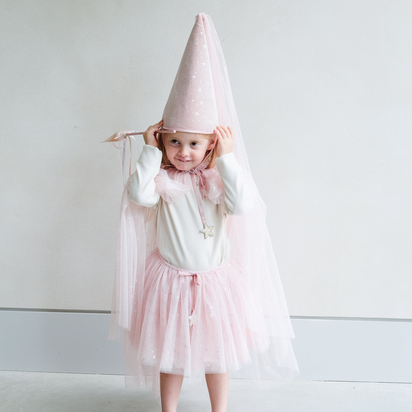Young girl wearing a full pink shimmer dressing up costume in pink with wand, princess hat, and matching tutu and cape against a white background