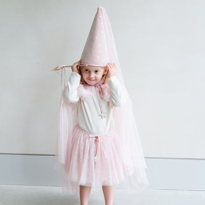 Young girl wearing a full pink shimmer dressing up costume in pink with wand, princess hat, and matching tutu and cape against a white background