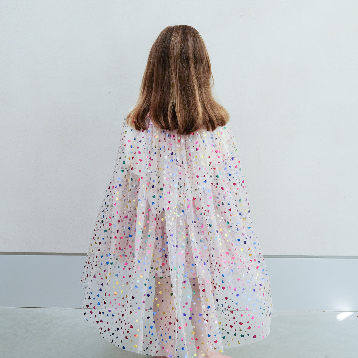 girl wearing rainbow heart cape and rainbow heart tutu against a plain background