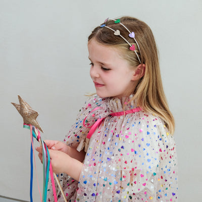 Young girl in a heart cape holding a star-shaped rainbow wand against a plain background