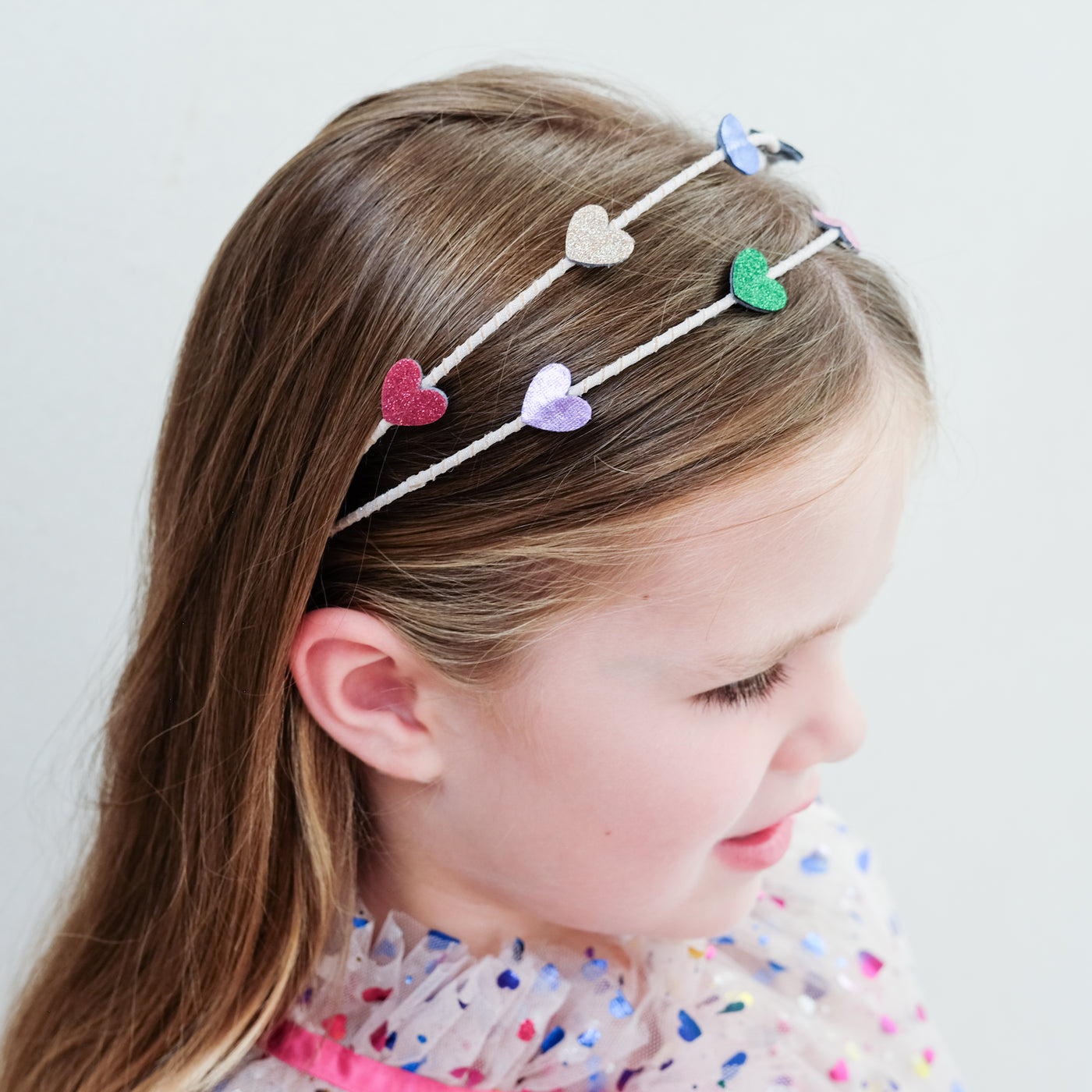 young girl wearing an rainbow heart alice band against a white background