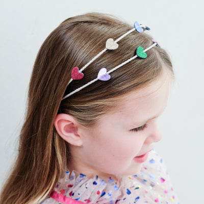 young girl wearing an rainbow heart alice band against a white background