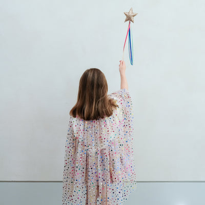 Young girl in a heart cape holding a star-shaped rainbow wand against a plain background