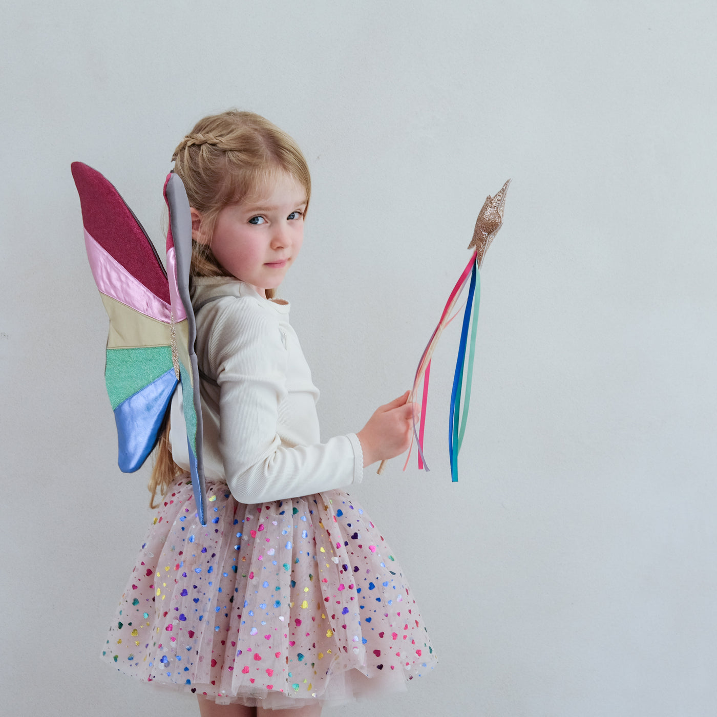 Little blonde girl wearing brightly coloured rainbow striped fairy wings, a multicoloured heart foil print tutu skirt and holding a gold glitter star wand with rainbow coloured ribbon tassels