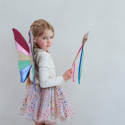 Little blonde girl wearing brightly coloured rainbow striped fairy wings, a multicoloured heart foil print tutu skirt and holding a gold glitter star wand with rainbow coloured ribbon tassels
