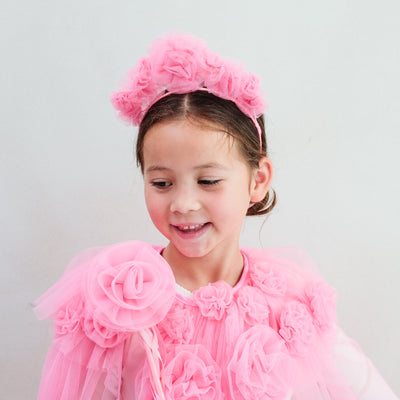 Young girl wearing a pink floral headband and pink cape against a light grey background
