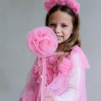 Young girl in a pink cape with floral headband and pink flower wand against a plain background