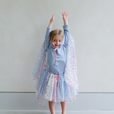Young girl wearing a colourful star-patterned rainbow cape and tutu against a plain background