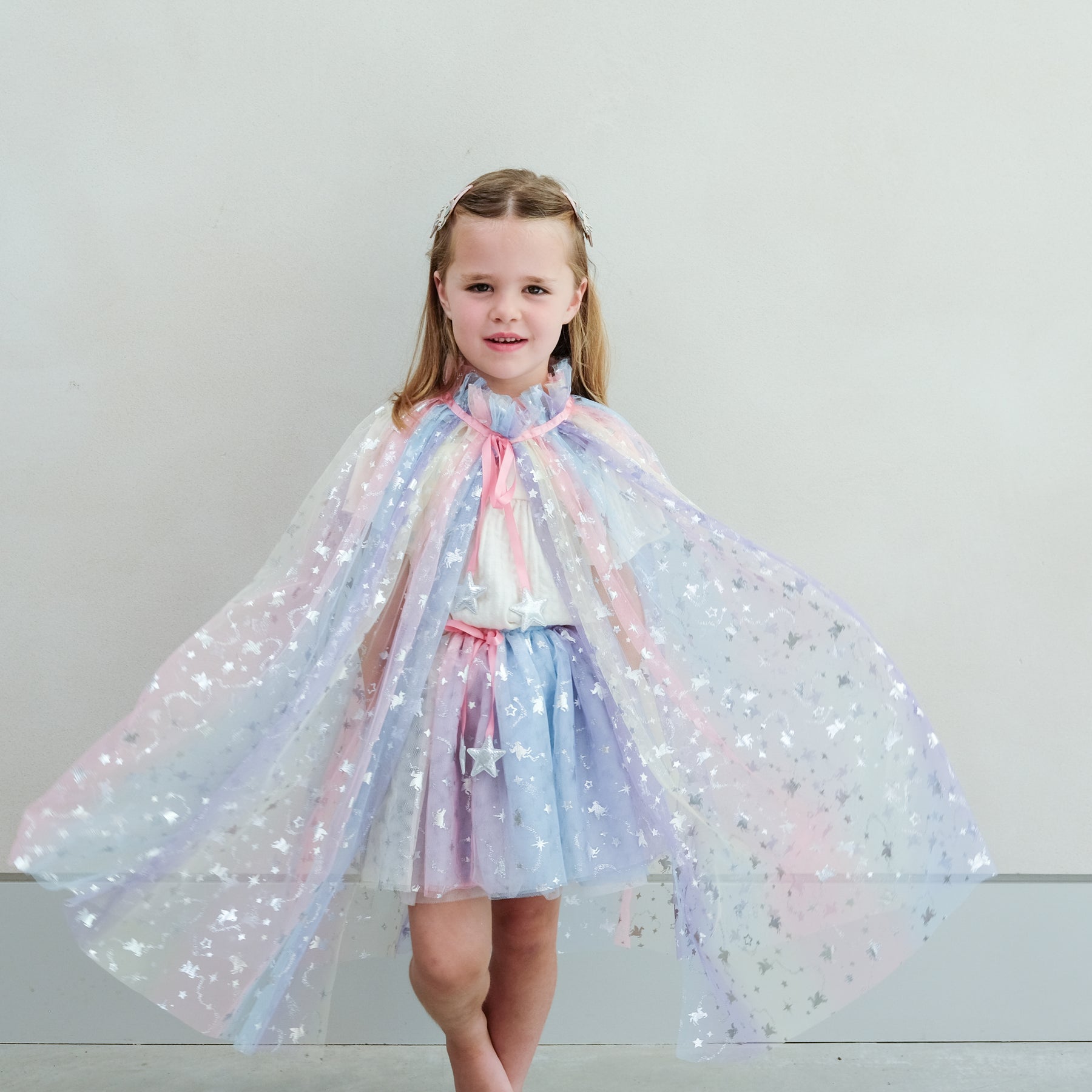 Young girl wearing a colourful star-patterned rainbow cape and tutu against a plain background
