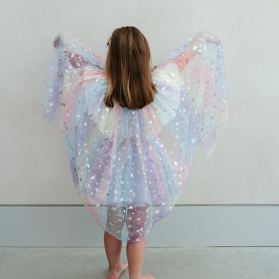 Young girl wearing a colourful star-patterned rainbow cape and tutu against a plain background