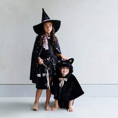 Two children in Halloween costumes, one in a witch outfit and the other in a black cat costume, standing against a plain background.