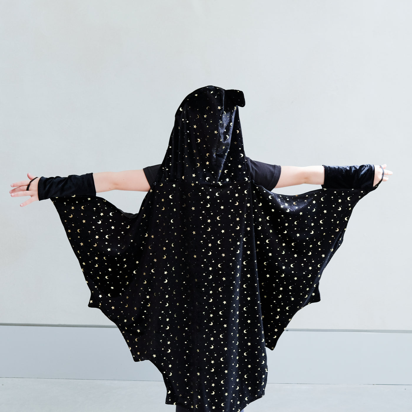 Child wearing a black hooded bat cape with star patterns against a grey background