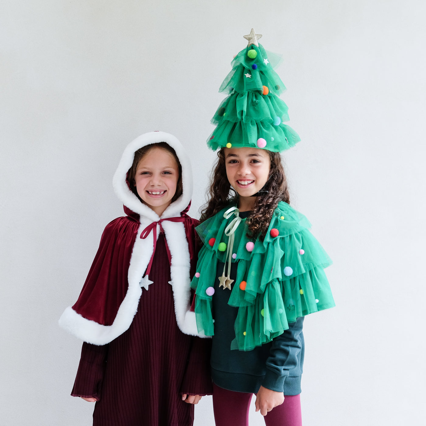 Two children wearing festive costumes, one resembling a Christmas tree and the other a Mrs Claus cape, on a plain background.