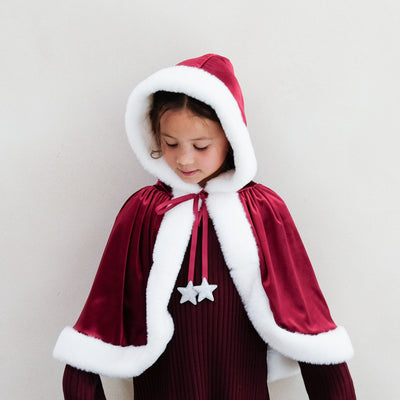 Child wearing a red and white hooded Mrs Claus cape with a star trim tie on a plain background