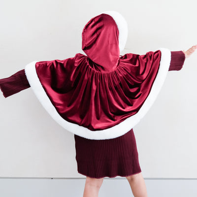 Child wearing a red and white hooded Mrs Claus cape with a star trim tie on a plain background