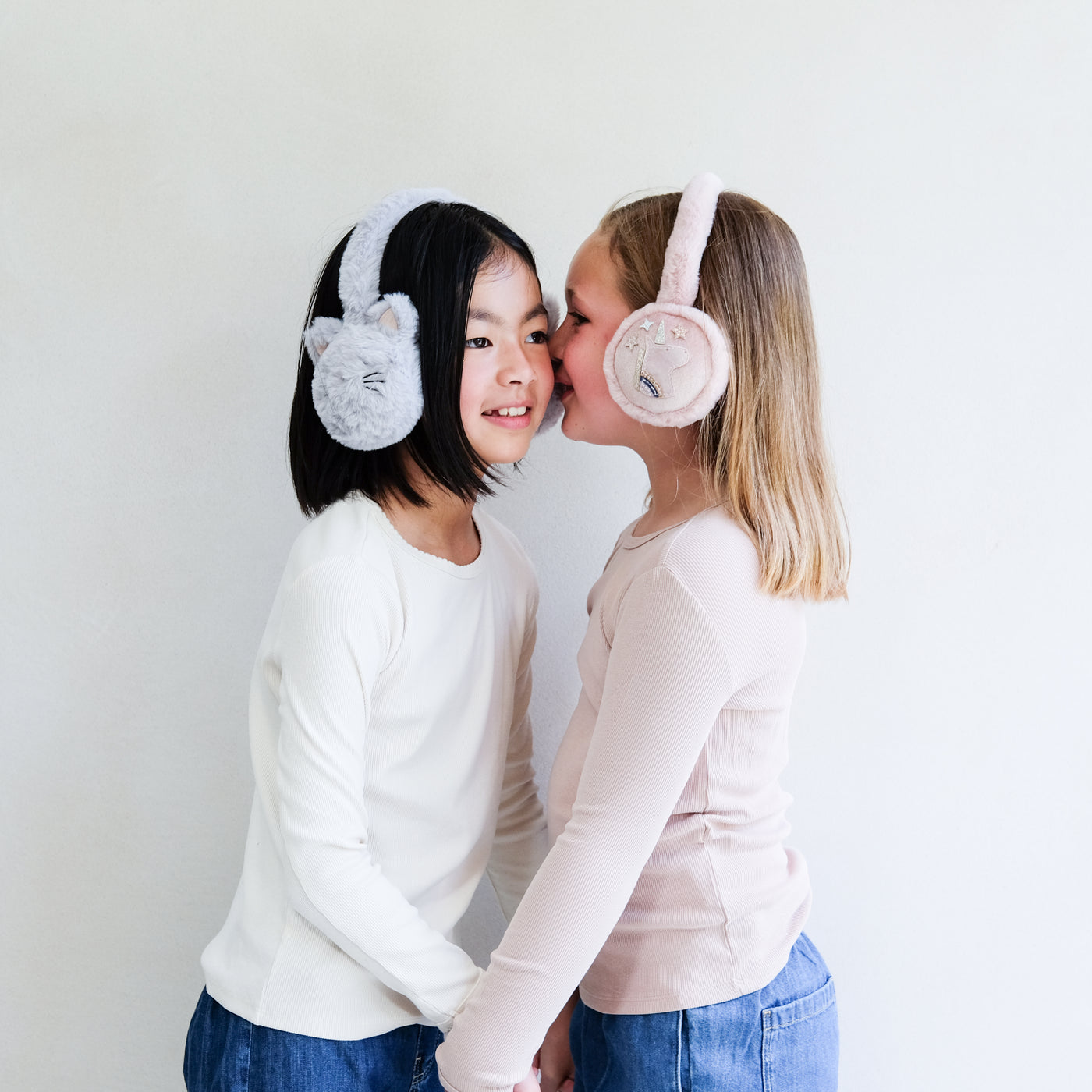 Two children wearing fluffy earmuffs, one with cat ears, the other with a unicorn and stars, standing close together against a plain background.