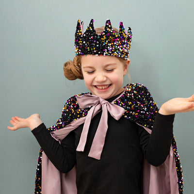 Little girl holding hands out wearing a rainbow sequin cape with large satin bow tie to the neck and matching sequin crown