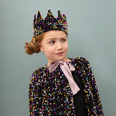 Little girl with alarmed look on green background with a side bun wearing a charming sequin covered cape and crown