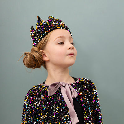 Little girl looking proud with shoulders back on a green background wearing fancy dress items including a beautiful sequin embellished cape and crown