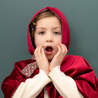 Little girl with a shocked face and hands up wearing a deep red velvet hooded cape