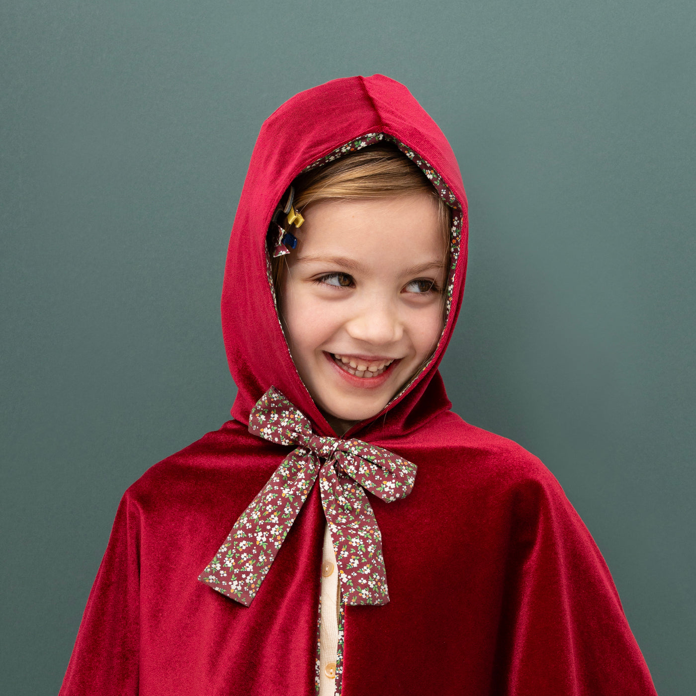 little girl smiling looking to side wearing a luxe velvet red hooded cape with floral bow