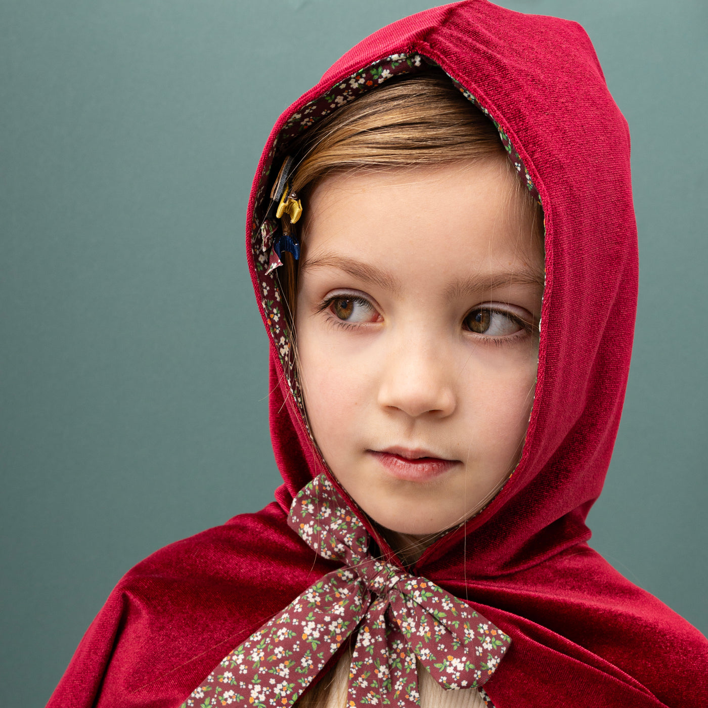 Close up of little girl with hazel eyes on a green background wearing a hooded red velvet cape with pretty floral bow tie