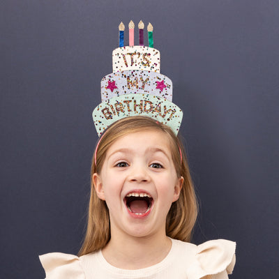 Little girl smiling and happy wearing a big birthday celebration headdress in the shape of a three tiered cake with candles made from glitter and metallic fabric