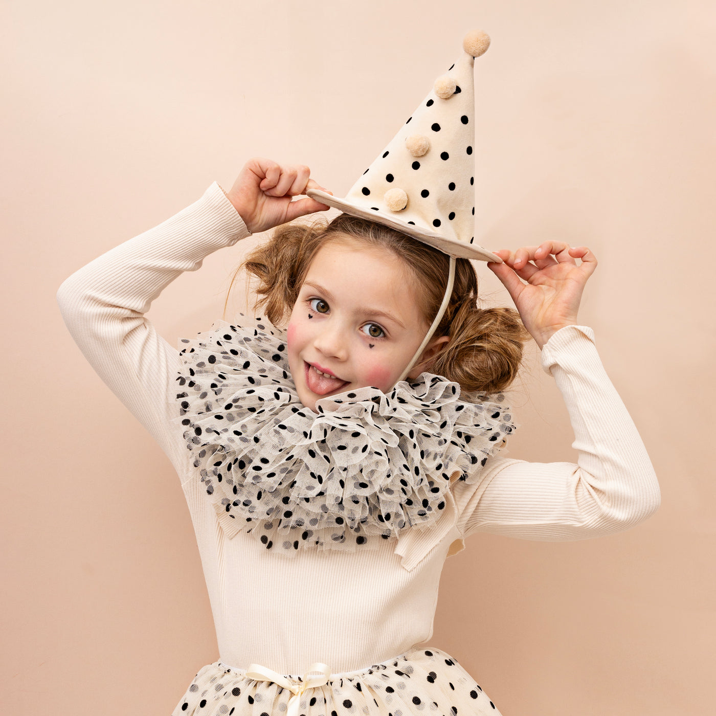 Little girl with hair in two buns wearing a polka dot clown hat with pom poms and a tulle ruffle clown collar and matching tulle tutu