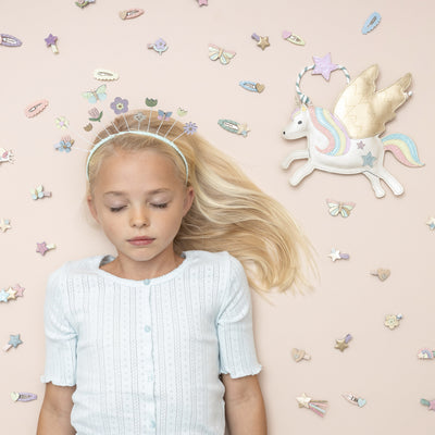 Little blonde girl with eyes closed surrounded by pretty pastel hair clips and a pearlescent fabric unicorn shaped bag, wearing a spring inspired flower and butterfly headdress