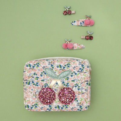 Zip top Floral fabric print bag featuring glittering cherries motif and rope strap on green background with cherry hair clips above