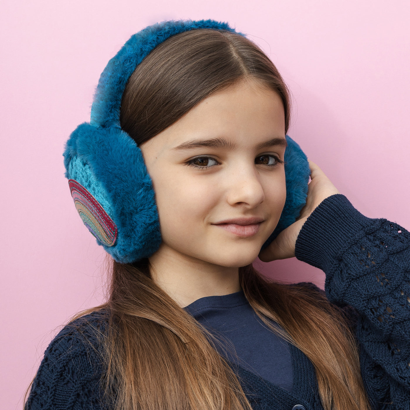 Dark haired girl smiling on pink background wearing colourful rainbow earmuffs with soft, fluffy padding, designed to keep little ears warm and cosy