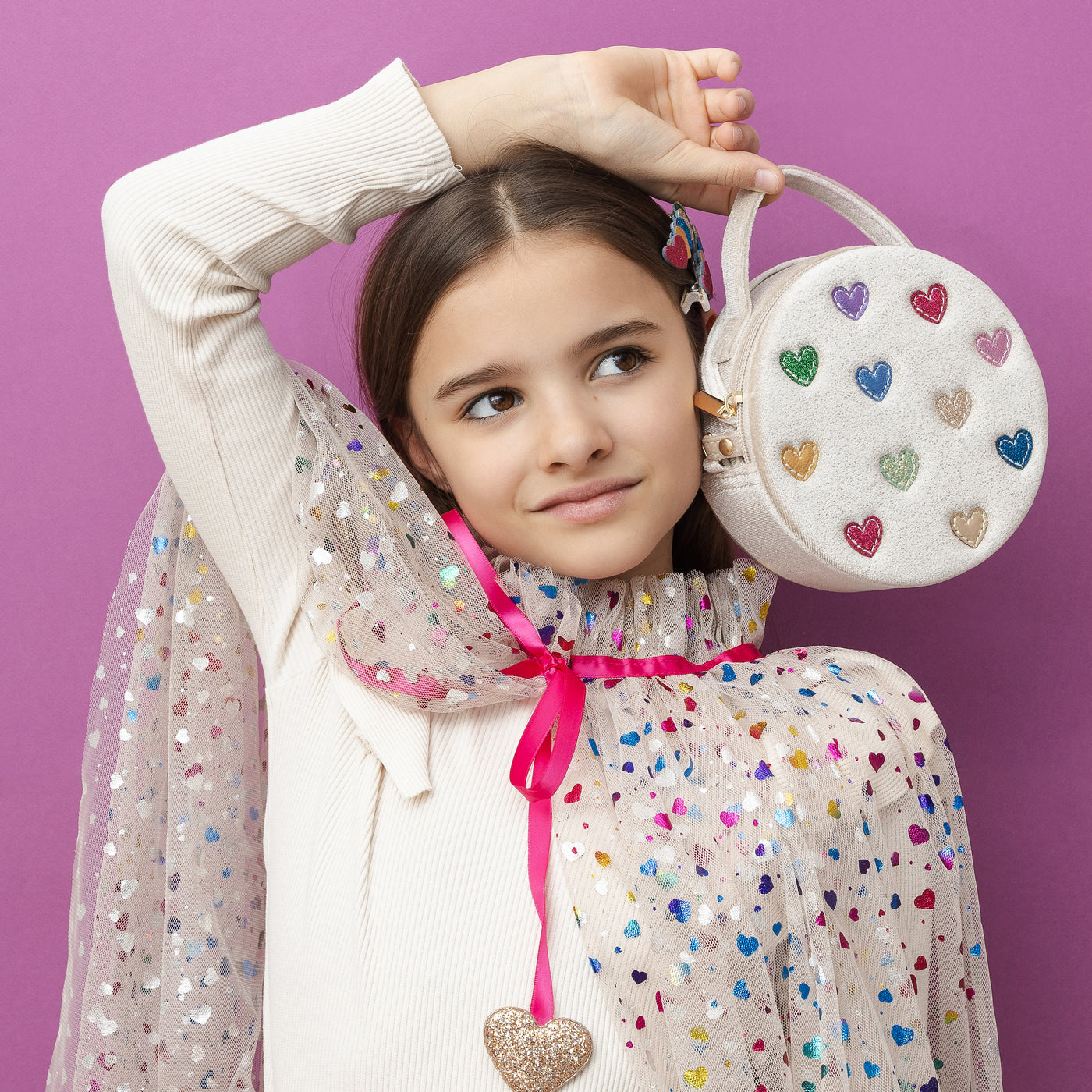 Dark haired girl on purple background holding a cream velvet jazzy heart bag- multi coloured hearts- zip closure- featuring a grab handle and a cross body strap, wearing a rainbow hearts cape and glitter heart hair clips